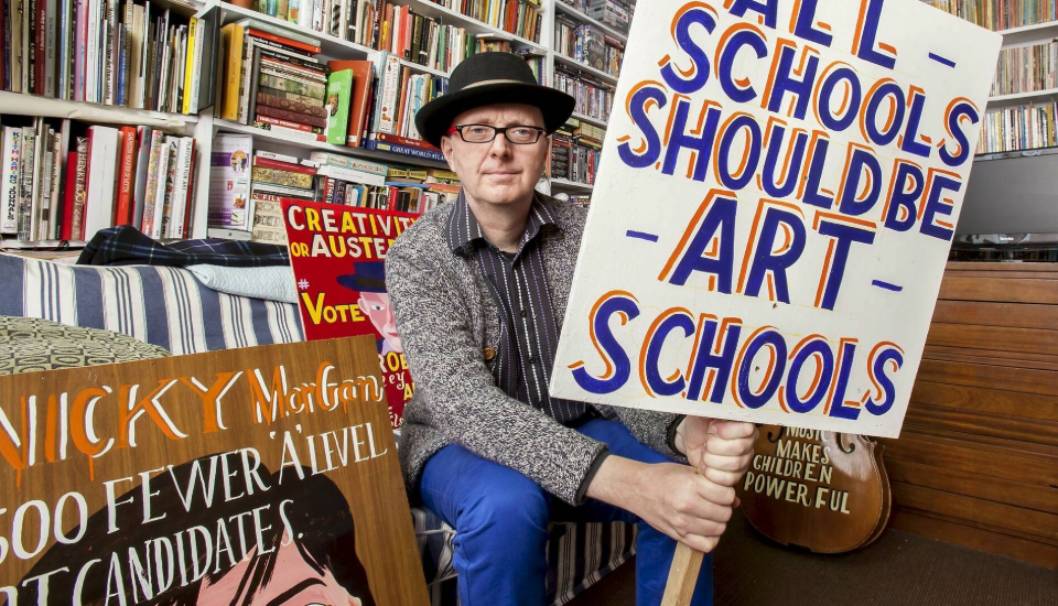 A person wearing glasses and a hat sits on a couch in a room full of books, holding a large sign that reads “ALL SCHOOLS SHOULD BE ART SCHOOLS.” Other colorful signs are visible around them.