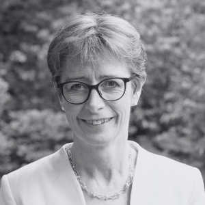 A woman with short hair and glasses, wearing a light-colored blazer and necklace, smiling at the camera. The background is outdoors with blurred greenery. The photo is in black and white.