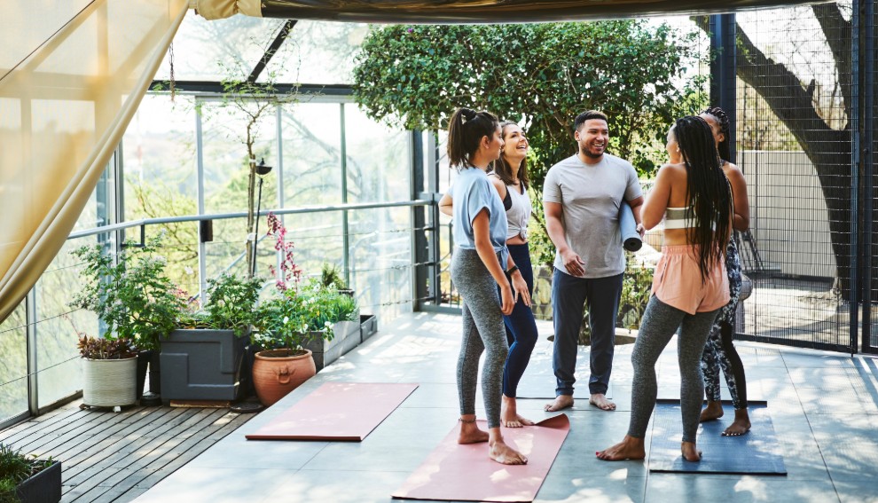 Five people stand and chat on yoga mats in a bright, open room with large windows and plants. Barefoot in athletic wear, they appear relaxed and happy, enjoying social connections as part of a group yoga or fitness class.