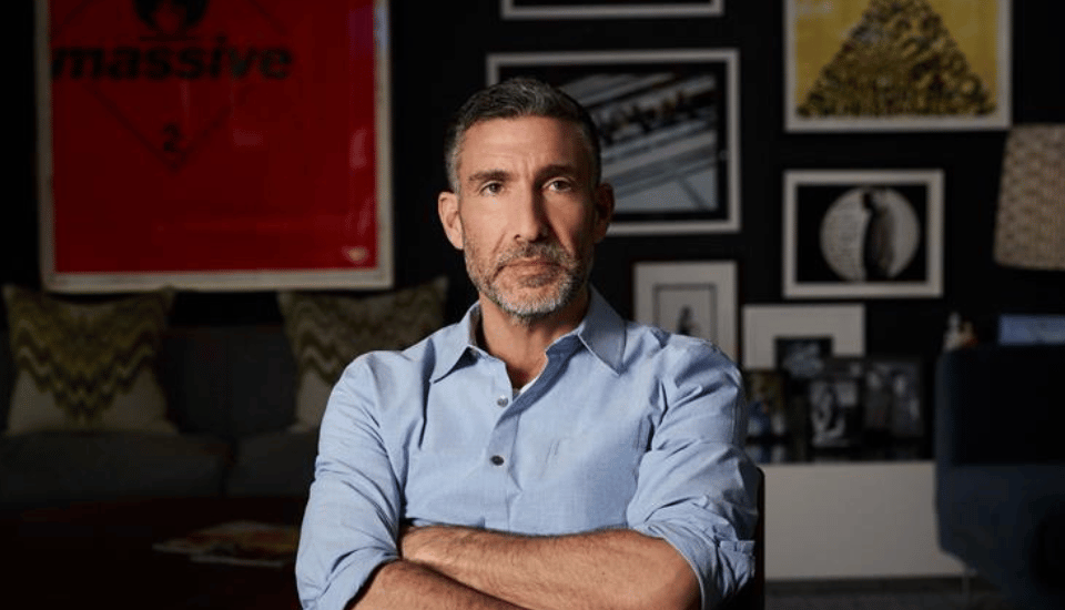 A man with short dark hair and a trimmed beard sits with his arms crossed, wearing a light blue shirt. He is indoors, in front of a dark wall decorated with framed artwork and photos.