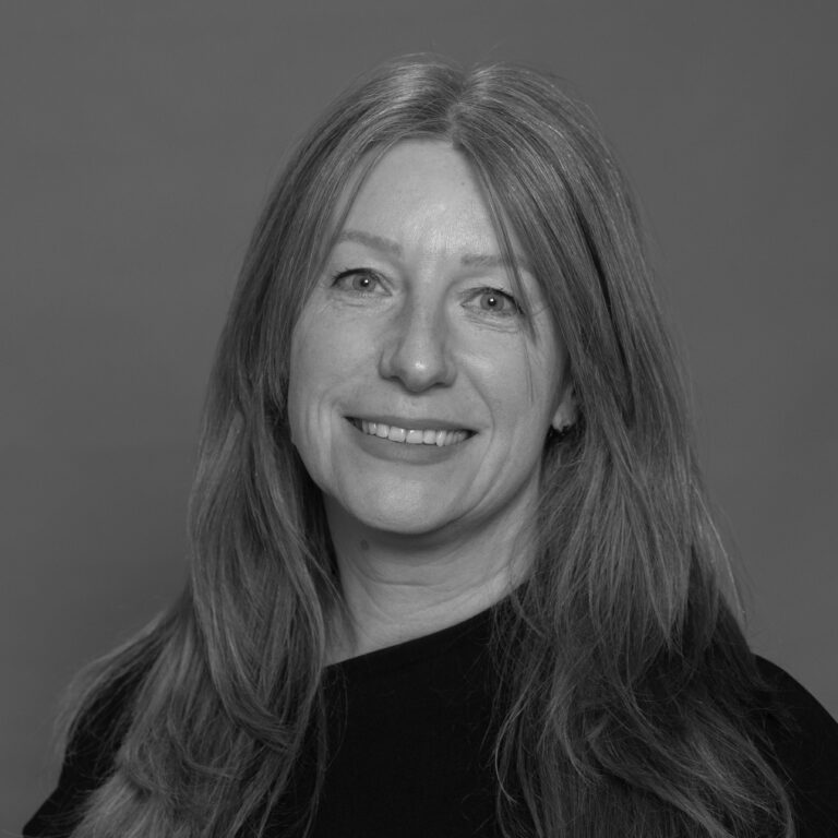 Black and white portrait of a smiling woman with long, straight hair, wearing a dark top, in front of a plain background.