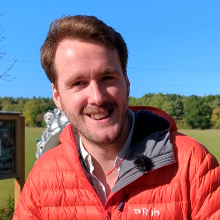 A man with light brown hair and a mustache smiles outdoors, wearing an orange jacket. Behind him are green trees, a blue sky, and an outdoor display sign partially visible on the left.
