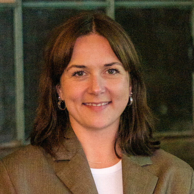 A woman with shoulder-length brown hair, wearing a brown blazer over a white top, smiles softly at the camera. The background is dark with blurred window panes.