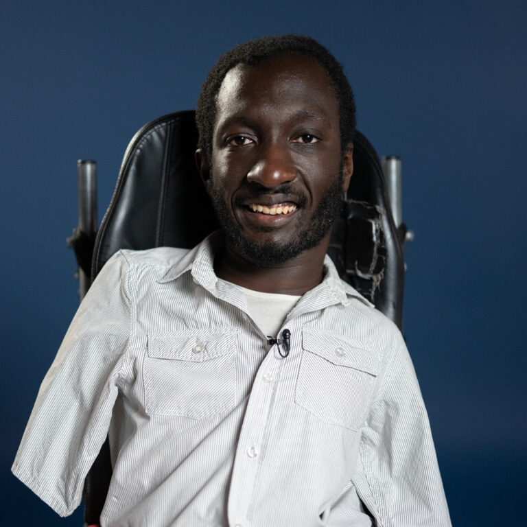 A man with short curly hair and a beard, wearing a light-colored button-up shirt, sits in a wheelchair against a plain dark blue background, smiling at the camera.