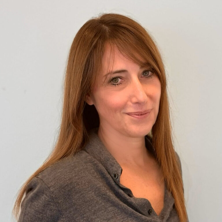 A woman with straight, long light brown hair and side-swept bangs is wearing a grey button-up top and smiling softly against a plain light-colored background.