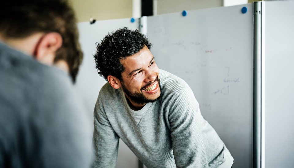 A man with curly hair and a beard, wearing a light gray sweater, leans forward and smiles brightly while discussing upcoming Hull events in an office setting. Whiteboards and another person are visible in the background.