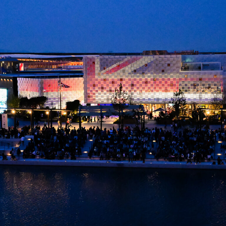 A large, modern building with a colorful, illuminated facade is seen at dusk. Crowds of people gather along a waterfront, and soft lights reflect on the water, creating a lively, festive atmosphere.