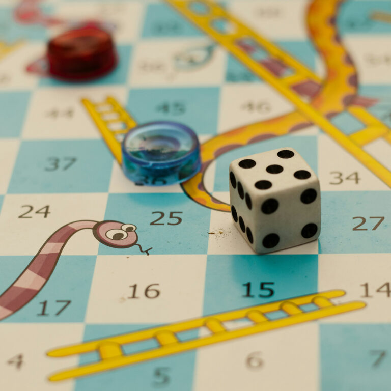A close-up of a Snakes and Ladders game board shows a six-sided die, two colored game pieces, a snake, and a yellow ladder on numbered squares.