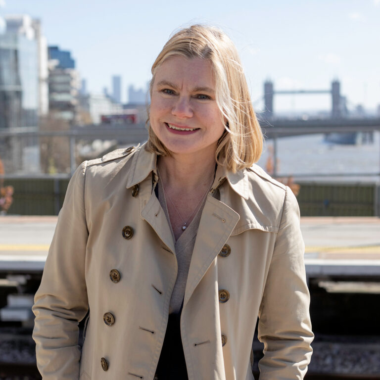 A woman with blonde hair in a beige trench coat smiles outdoors with blurred city buildings and a bridge visible in the background.