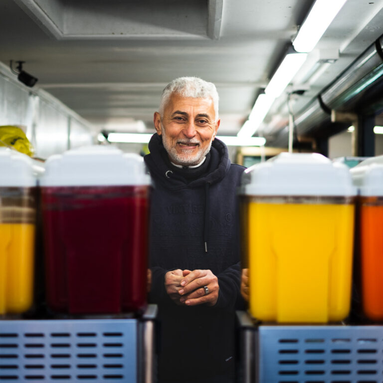 A smiling man with gray hair stands behind large beverage dispensers filled with colorful drinks in a brightly lit indoor setting.