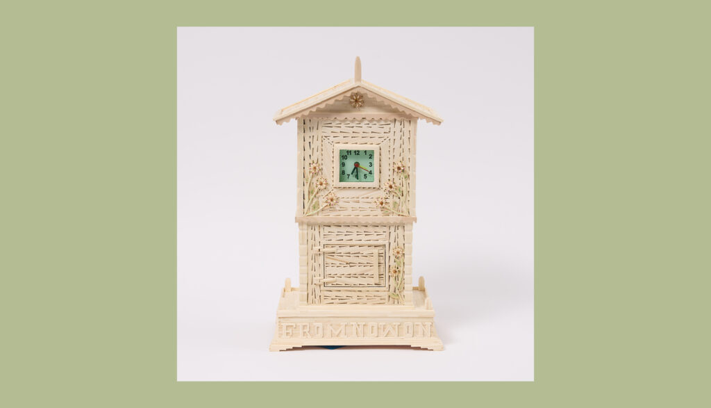 A small, cream-colored decorative clock shaped like a house, with intricate wicker-like patterns and floral details, is displayed against a plain white background with a pale green border.