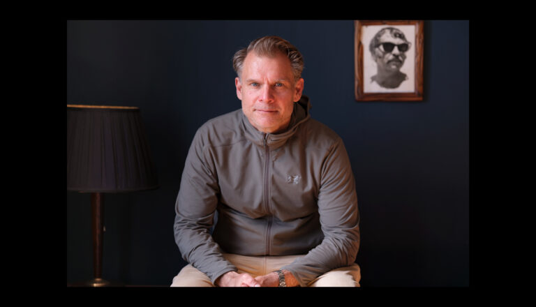 A man wearing a gray zip-up jacket sits in front of a dark blue wall, looking at the camera. A black floor lamp and a framed portrait of a man with sunglasses are in the background.