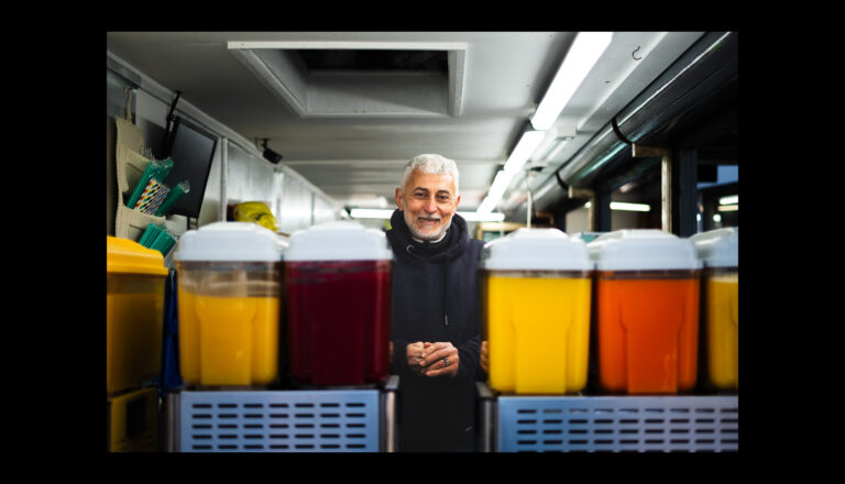 A smiling man stands behind a counter with large transparent containers filled with colorful juices in a well-lit indoor market or shop.