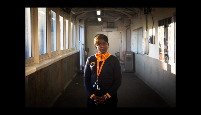 A woman in a navy uniform with orange accents stands in a sunlit corridor, facing the camera with keys hanging from her waist. The corridor has windows on one side and posters and signs on the walls.