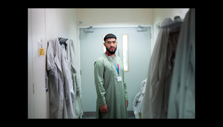 A man wearing a long green robe and a lanyard stands in a hallway lined with hanging garments, looking toward the camera. Light comes through a door with frosted glass behind him.