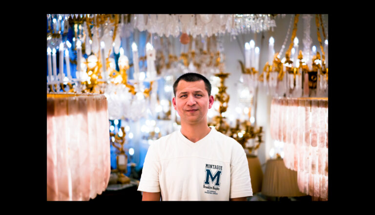 A man in a white T-shirt stands indoors surrounded by hanging chandeliers and decorative lights, with a soft, warm glow illuminating the scene.