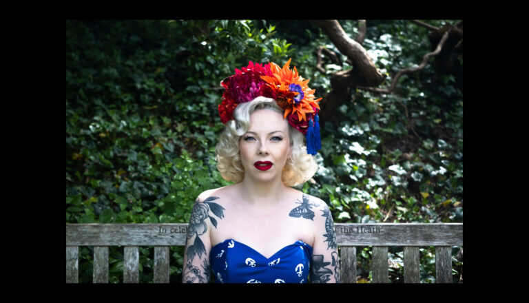 A woman with curled blonde hair, bright red lipstick, tattoos, and a strapless blue dress sits on a wooden bench. She wears a colorful flower headpiece. Lush green foliage and tree branches fill the background.