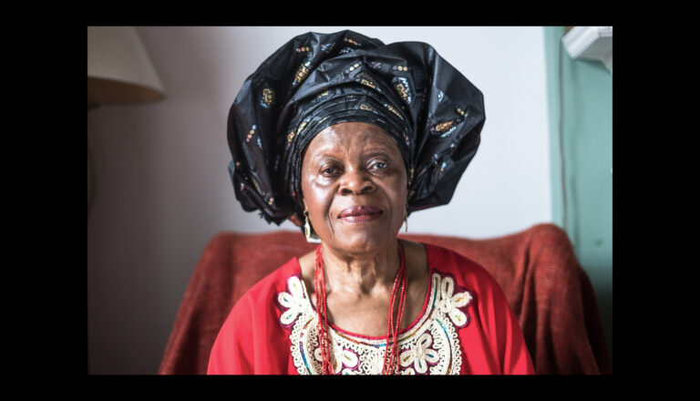 An elderly woman wearing a black, patterned headwrap and a red, embroidered dress sits indoors, looking at the camera with a gentle expression. A red cloth covers the chair behind her.