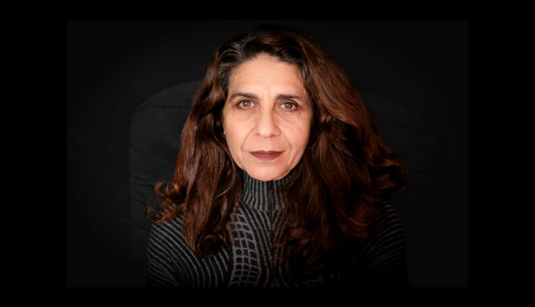 A woman with long, wavy brown hair and a serious expression looks directly at the camera. She is wearing a dark, patterned turtleneck sweater and sits against a plain black background.