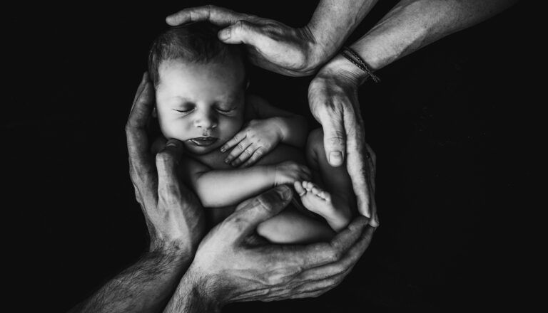 Black and white photo of a sleeping newborn cradled in adult hands, forming a protective circle around the baby, symbolizing care, safety, and love against a dark background.