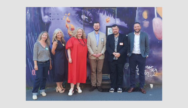 Six people stand in a row, smiling, in front of a colorful street mural featuring a large face. They are dressed in smart-casual attire and appear to be attending an event or gathering outdoors.