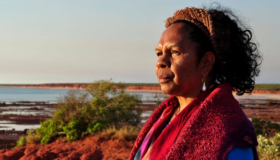 A woman with curly hair, wearing a headband, dangly earrings, and a red shawl, stands outdoors near a rocky coastline, gazing thoughtfully into the distance at sunset.
