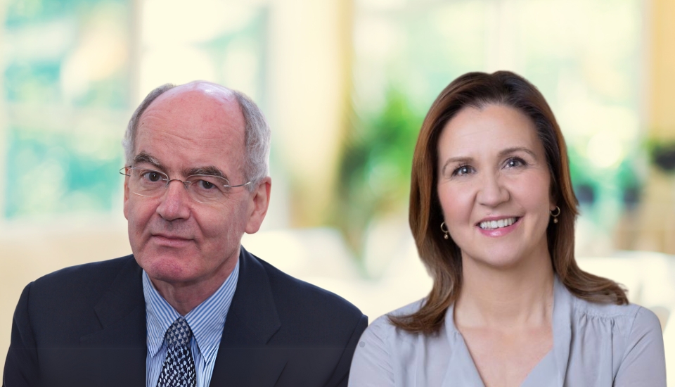 A man in a suit and glasses sits next to a smiling woman with brown hair, both posed in a bright, softly blurred indoor setting.