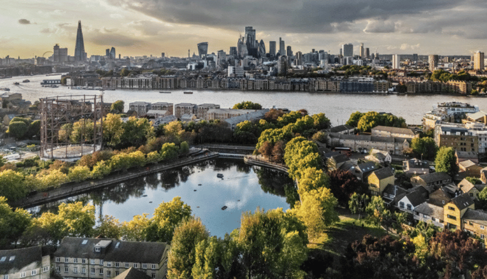 A panoramic view of London featuring the River Thames, lush green parks, residential buildings, and the city skyline with landmarks like The Shard and modern skyscrapers under a cloudy sky.