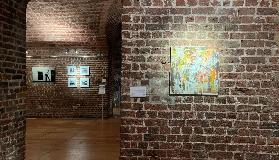 A brick-walled art gallery with wooden floors displays abstract and black-and-white artworks; one colorful abstract painting is featured on the right wall, and framed photographs hang on the far wall.