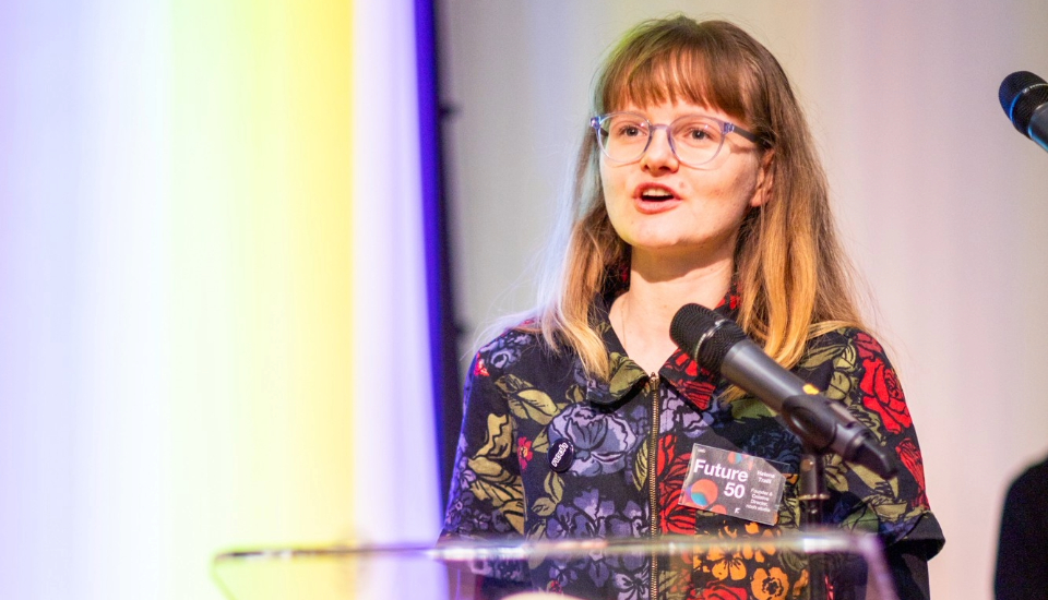 A person with long hair and glasses speaks at a podium with a microphone. They wear a colorful floral-patterned shirt and a “Future 50” badge. A blurred background features yellow and purple lighting.