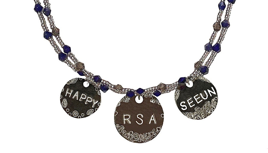 A necklace with three silver circular pendants, each engraved with patterns and letters. The left says "HAPPY," the middle has "RSA," and the right has "SEEUN." The chain is decorated with blue and silver beads.