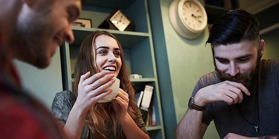 Three young adults sit together at a table. One woman smiles while holding a cup, while the two men next to her are engaged in conversation. Bookshelves and a large wall clock are visible in the background.