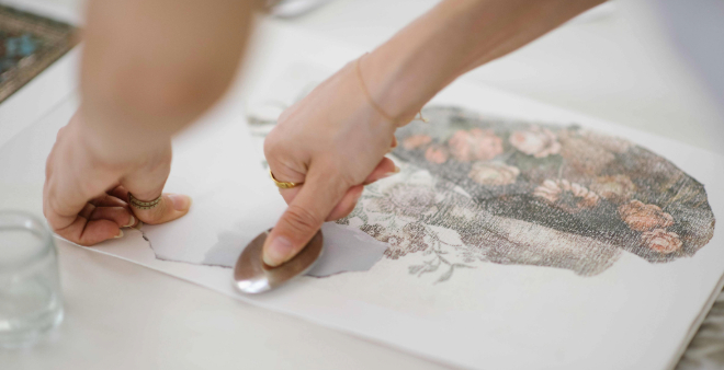 Close-up of hands transferring a floral image onto paper using a metal spoon, with a glass jar nearby on the table.