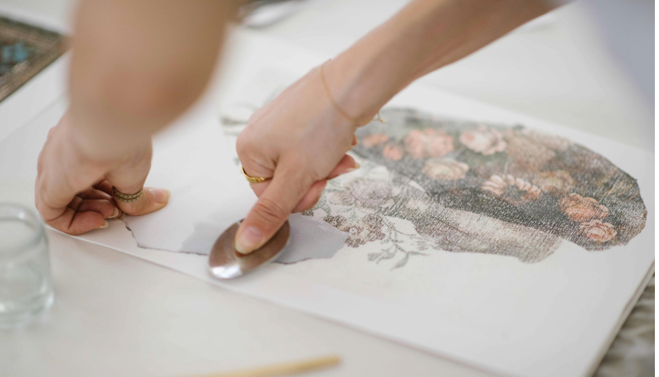 A person uses a metal tool to transfer or smooth an image of a floral arrangement onto a white surface, likely as part of an art or craft project.