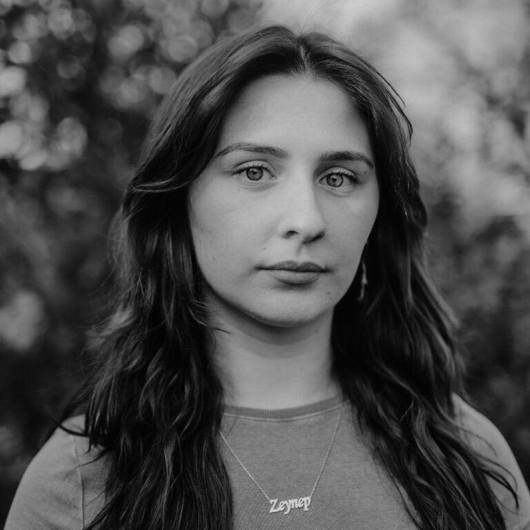 Black and white portrait of a young woman with long, dark hair, wearing a necklace that says "Zeynep." She has a neutral expression and stands outdoors, suggesting a quiet connection to the blurred natural background.