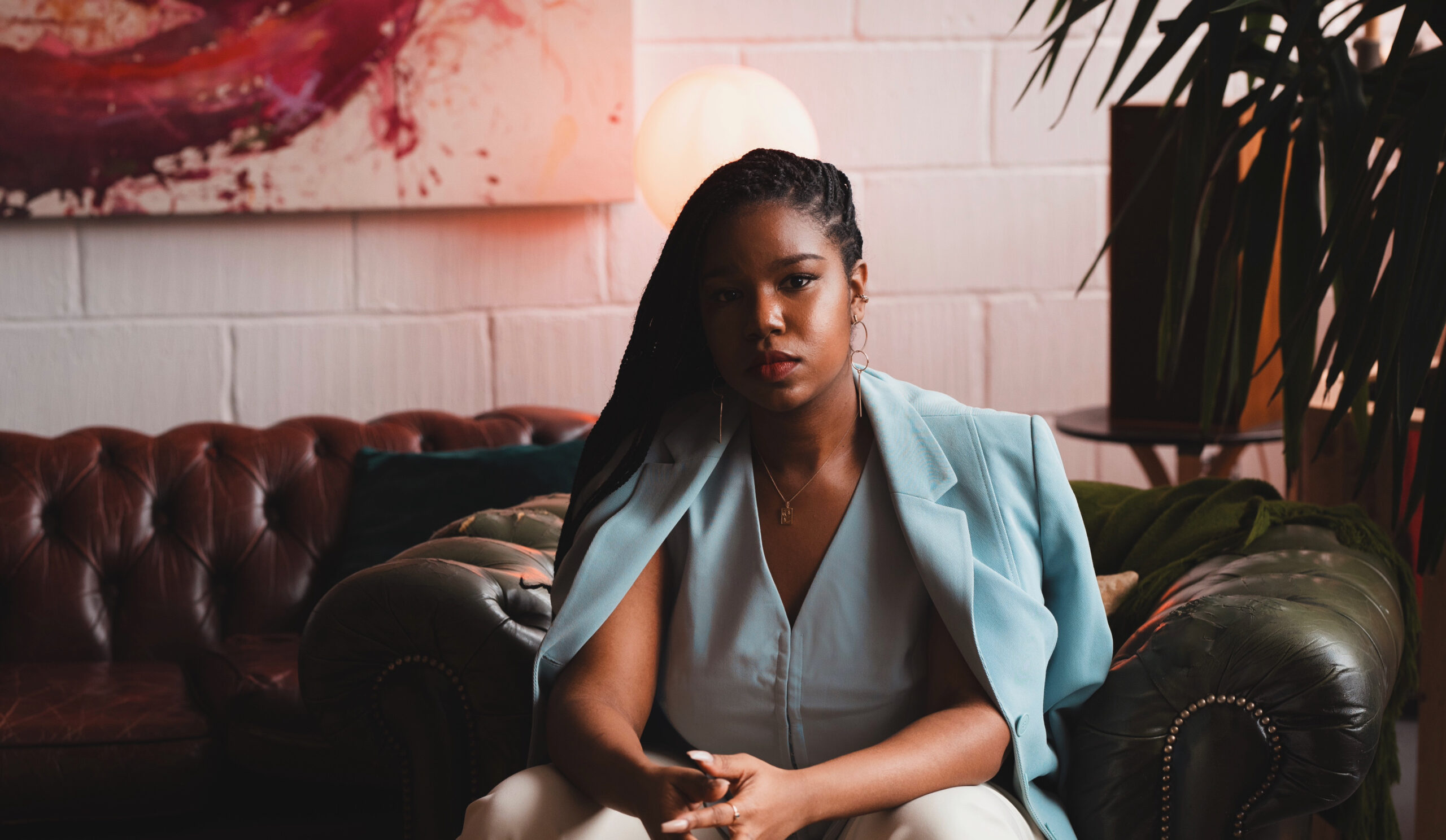 A woman with long braided hair sits on a leather couch, wearing a light blue blazer draped over her shoulders. She looks thoughtful, as if preparing for a leadership workshop London, in a stylish room with modern art and soft lighting.