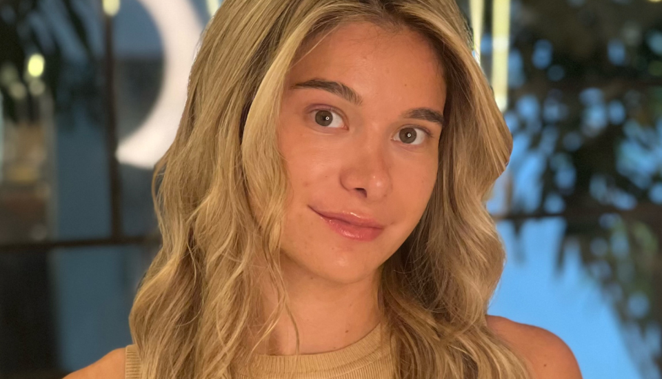 A young woman with long, wavy blonde hair and a tan sleeveless top stands indoors, looking at the camera with a slight smile, capturing an approachable style reminiscent of Lynsey Shaw FRSA. The softly blurred background hints at plants and lights.