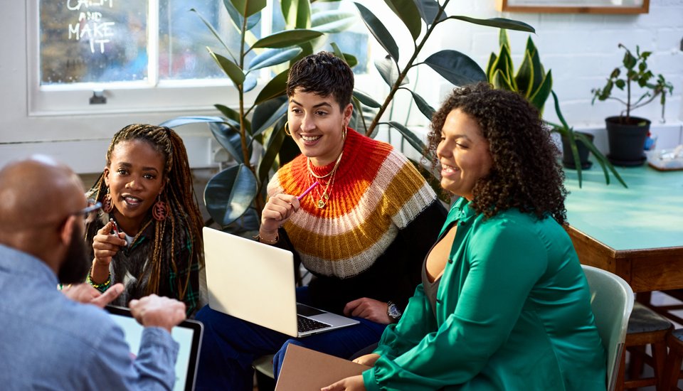 Four people sit together in a brightly lit room with plants, engaged in conversation about autumn events. Two have laptops open, and everyone appears focused and cheerful as they plan upcoming gatherings.