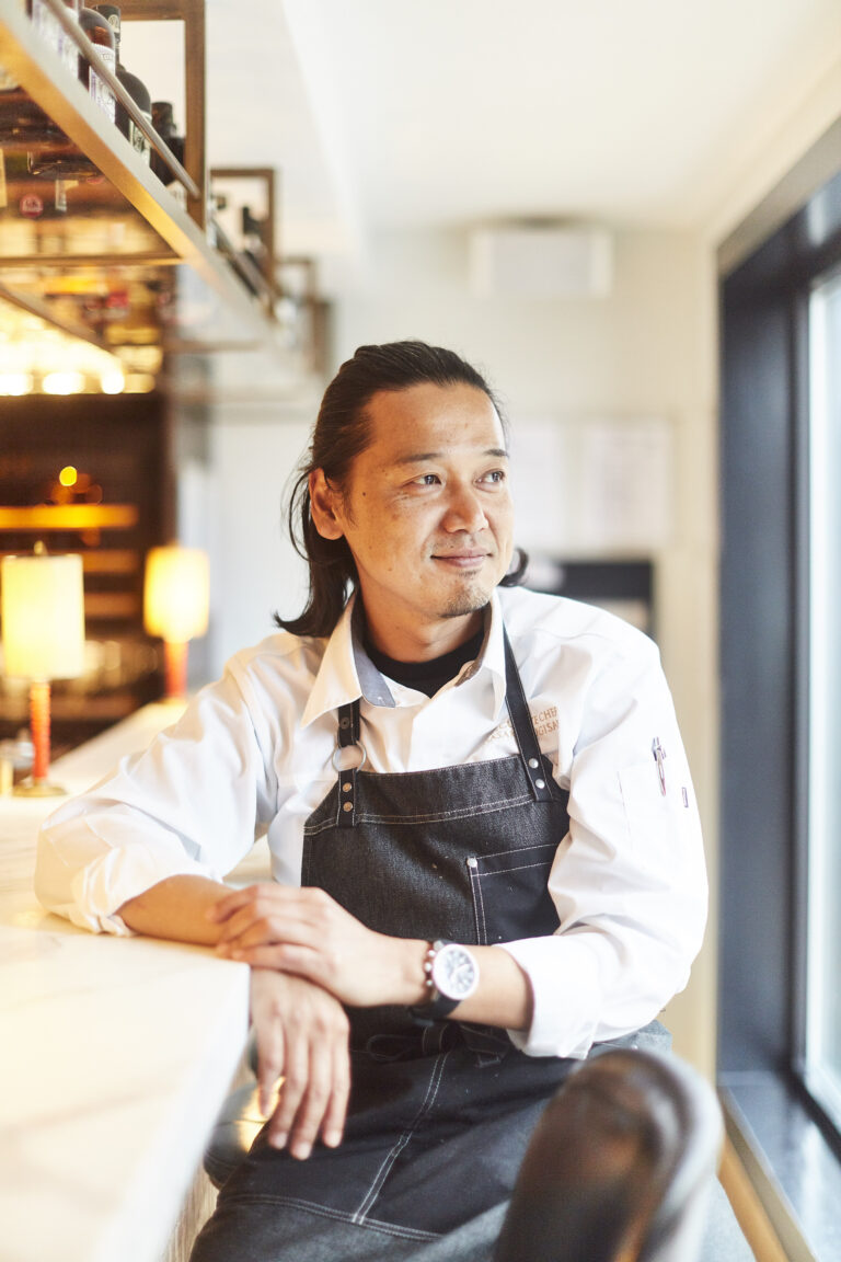 A chef wearing a white shirt and dark apron sits at a counter, smiling and looking to the side in a bright, modern restaurant setting.