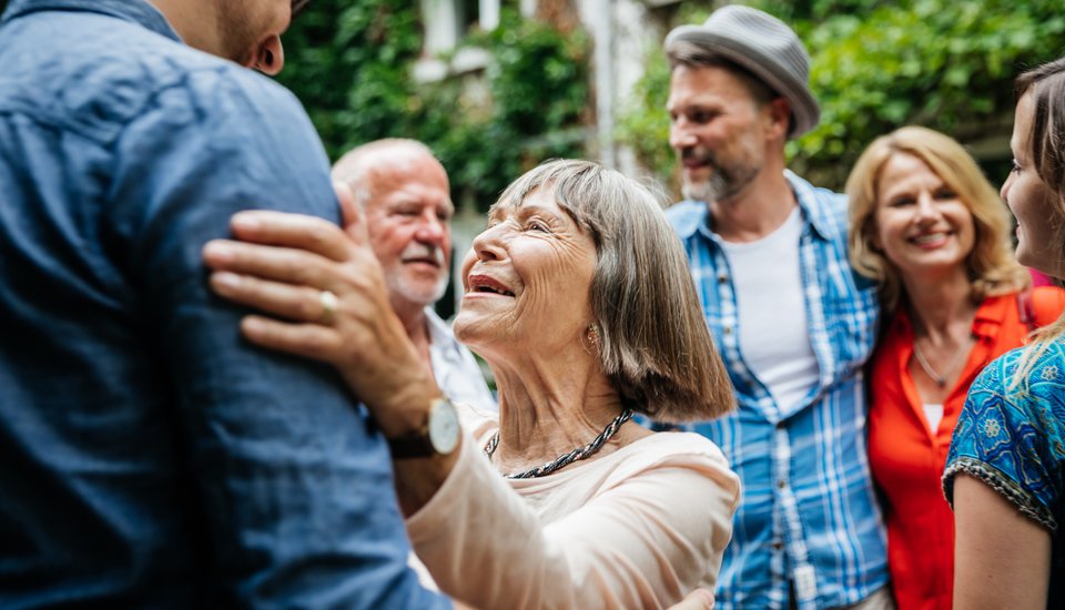 An elderly woman smiling warmly and embracing a man while surrounded by a group of people outdoors, all appearing happy and engaged with each other.