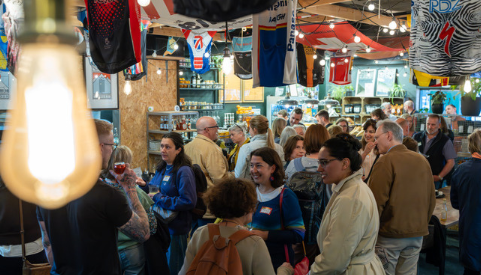 A crowded indoor social event with people chatting and mingling under hanging sports jerseys and string lights, with a bright lightbulb in the foreground. Shelves and displays are visible in the background.