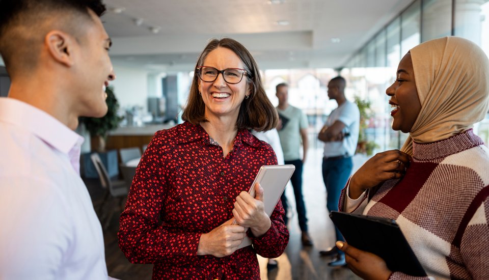 Three people stand and talk cheerfully in a modern office setting, one holding a tablet and another holding a notebook, with more people conversing in the blurred background.