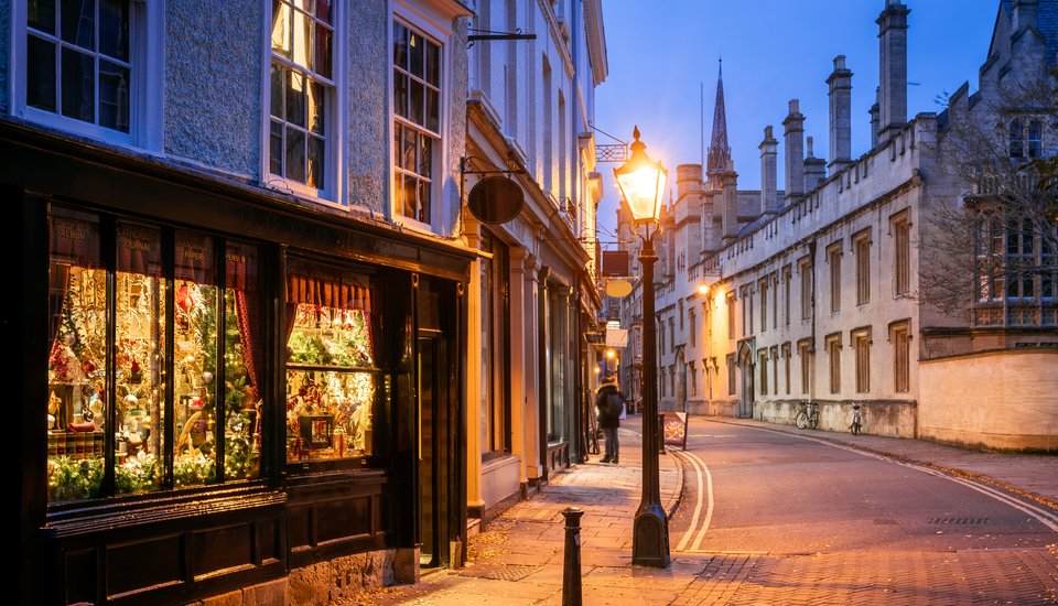 A warmly lit shop window decorated for Christmas glows on a quiet, cobblestone street at dusk, with historic stone buildings and a church spire in the background under a deep blue sky.