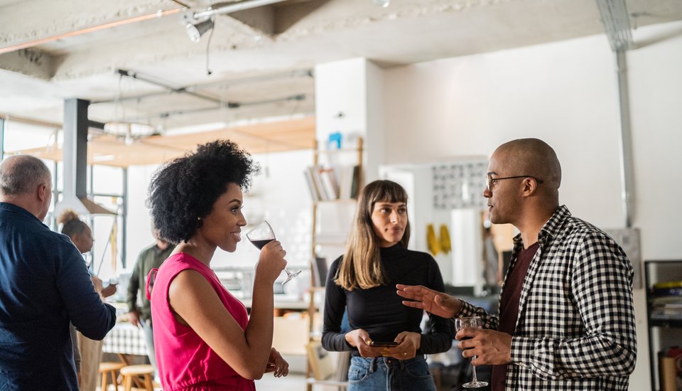 At a creative workshop, people socialize at a casual indoor gathering. A woman in a pink top sips from a wine glass while talking to a man in glasses and a checkered shirt, as another woman stands nearby listening.