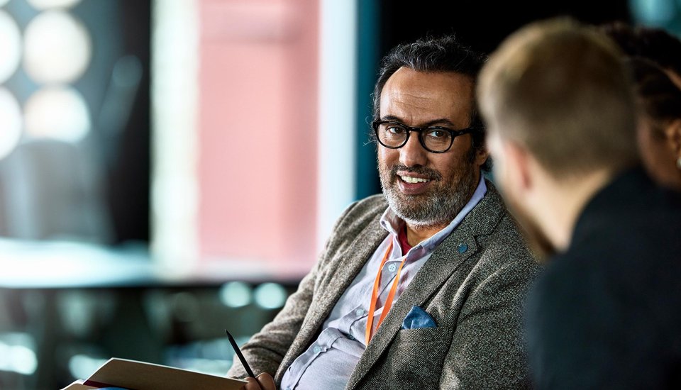 A man with glasses and a gray beard, wearing a blazer and orange lanyard, smiles while talking to another person in a blurred indoor setting.