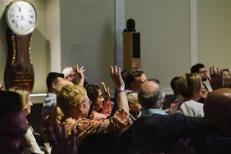 A group of people seated in a room raise their hands, participating in an event or meeting. A large clock is visible on the wall in the background. The atmosphere appears engaged and attentive.