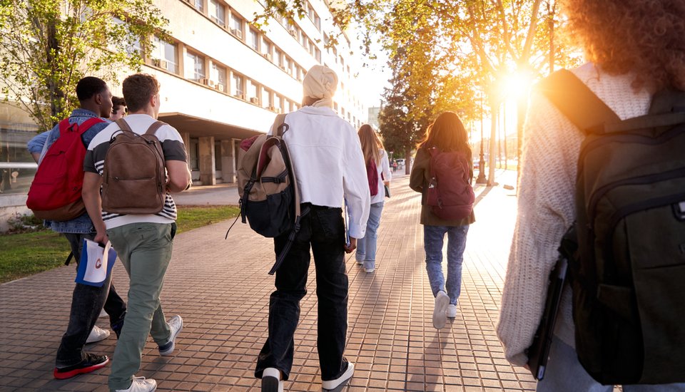A group of students with backpacks walk together on a tree-lined sidewalk in the sunlight outside a building, heading toward the campus entrance—ready to learn and shape the future of healthcare innovation.