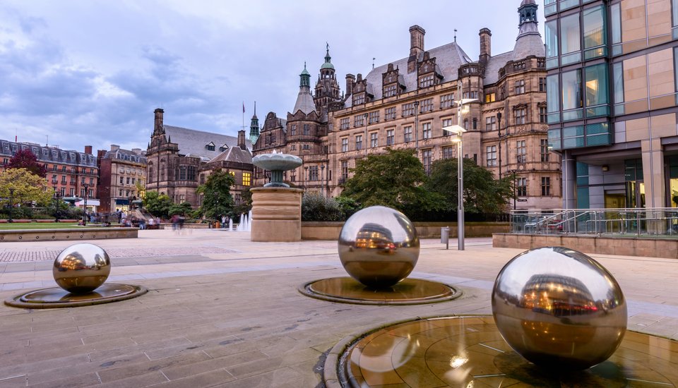 Reflective steel spheres and a fountain in a plaza with historic and modern buildings around, under a cloudy sky in Sheffield Peace Gardens, England—the perfect setting for an inspiring creative workshop.