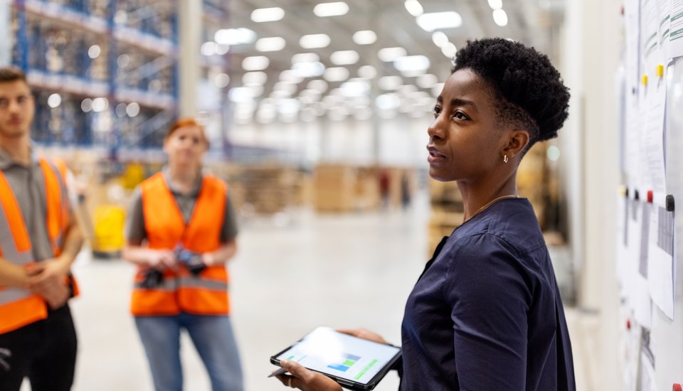 A woman holding a tablet stands by a whiteboard in a warehouse, addressing two people in orange safety vests who are listening attentively. Shelves and boxes are visible in the background.