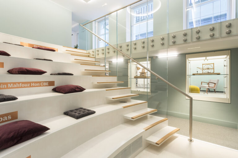 Modern indoor seating area with white tiered steps, each with dark red and gray cushions. Glass railing and large interior windows reveal more seating and a decorative clock in the next room. Bright and well-lit space.
